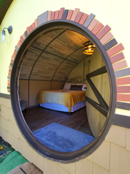 A round doorway leads into a cozy, rustic bedroom with a bed and warm lighting, surrounded by yellow walls.